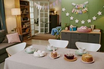 Fototapeta premium Birthday party table setting featuring cake with number three candle, pastries, teacups, and plates arranged in foreground, festive decorations and gifts visible in background