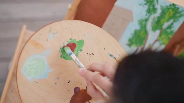 closeup palette mixing red paint hand applying brush, painter blending warm pigments into floral sketch on canvas, outdoor deck setting with natural light, quick dab technique and playful creative
