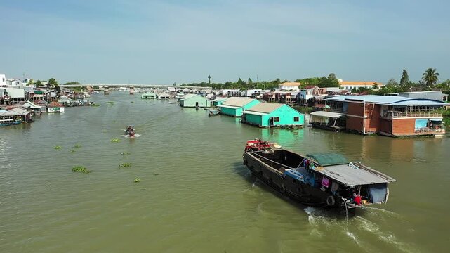 Aerial view of boats and colorful floating houses on the river in Chau Doc, Vietnam, under a clear sky. Traditional river life and vibrant waterfront structures are visible.