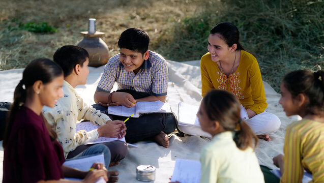 Indian villager girl boy sitting on mat do study read book outdoor rural area ground place. Happy group child hold notes talk gossip enjoy desi life new day outside open class field
