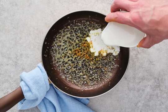 Cooking onions and spices in a pan while adding cream in a kitchen setting during daytime hours