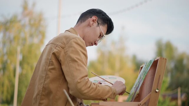 young asian artist painting on easel in tranquil autumn park, art student sketching foliage, landscape painter refining greens, illustrator mixing subtle tones, side profile concentration, casual