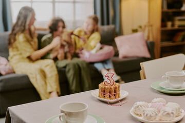 Fototapeta premium Caucasian woman, Caucasian teenager, and Caucasian child sitting on sofa interacting with small dog, birthday cake with number three candle and desserts arranged on table in foreground