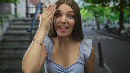 Fototapeta premium Caucasian teenage girl in blue top covers eyes with hand while smiling on sunlit urban street; shyness mystery.
