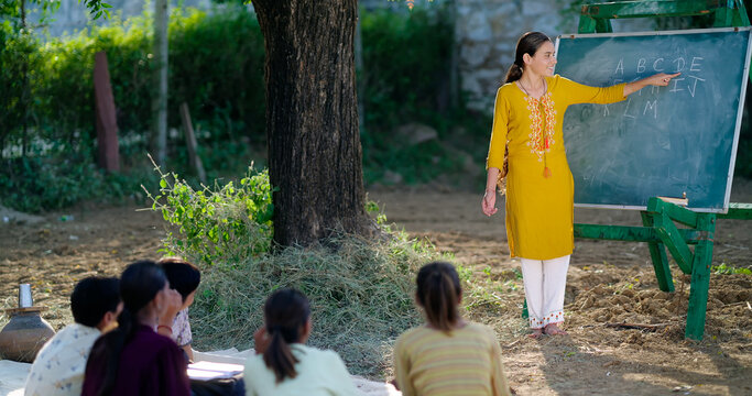 Indian Young adult gen z female tutor standing teach English to villager girl boy outdoor rural area. Happy child group sitting on mat look board learn do study new day time outside ground place