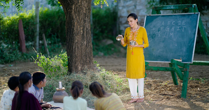 Indian happy female teach boy group do study sitting on mat rural area young girl show tiffin box in hand talk speak gossip enjoy new day desi life spend day break time outdoor open class air field
