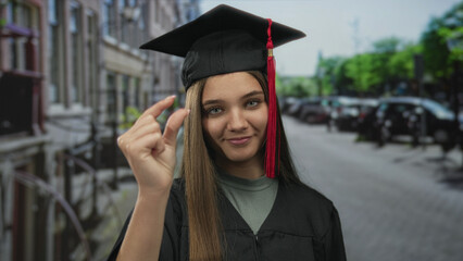 Fototapeta premium Young woman wearing graduation cap and gown pinches her fingers in front of blurred city street buildings and parked cars; pride.