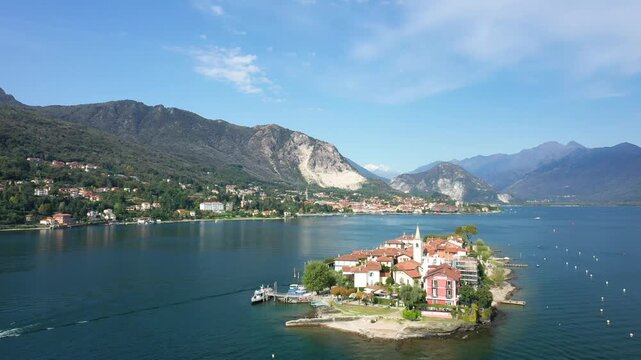 Aerial view of Isola dei Pescatori on Lake Maggiore with mountains in the background under a clear blue sky. Picturesque village and serene waters create a scenic landscape.