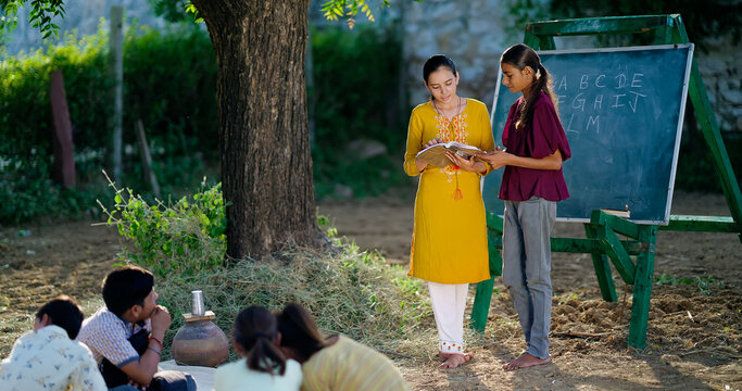 Indian young adult female hold book do study helping to read teen age girl class rural area place. Boy student group talk speak sitting on mat enjoy spend break time have fun joy open air ground