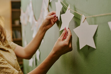 Fototapeta premium Caucasian young adult woman decorating wall by hanging paper star garland, hands carefully arranging decorations on string, preparing festive display against green background