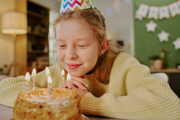 Fototapeta premium Caucasian girl child wearing party hat smiling and gazing at birthday cake with lit candles, resting chin on hands, celebrating birthday indoors with festive decorations in background