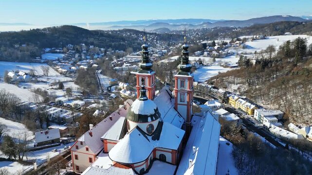 Aerial view of the snow-covered Basilica Mariatrost, standing majestically against the backdrop of rolling hills and a clear blue sky, Graz, Styria, Austria.