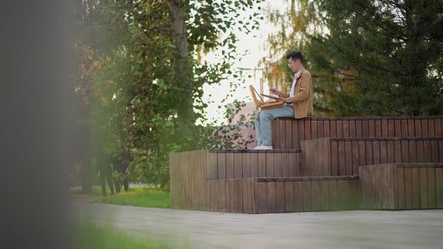 illustrator painting on tiered wooden benches golden hour, sunlight filtering through leaves with watercolor set and brush in hand, wearing jeans and jacket with focused posture, city path