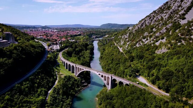 Aerial view of the bridges of Solkan spanning the turquoise river, contrasting with the lush green trees and the distant terracotta rooftops, Solkan, Slovenia.