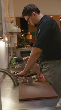 Professional chef opening chef knife roll and taking out tools on stainless steel counter while his colleague preparing ingredients for cooking in restaurant kitchen. Vertical video