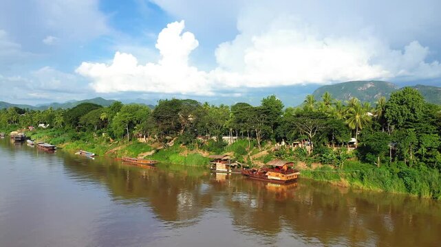 Scenic view of lush green trees and traditional wooden boats along a calm riverbank in Luang Prabang, Laos, with mountains in the background under a partly cloudy sky.