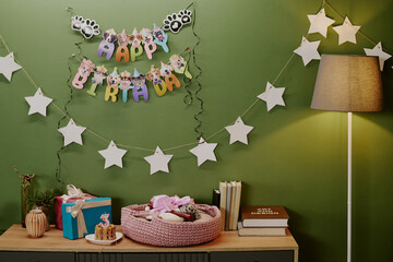 Fototapeta premium Birthday party setup featuring pink basket with stuffed animal, cupcake with number three candle, wrapped gifts, star garland, books, lamp, animal-themed happy birthday banner on green wall