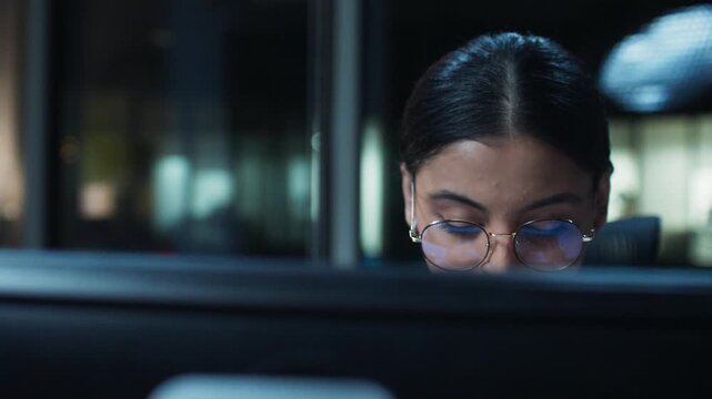 Close face view of businesswoman wearing glasses behind monitor. Adult female analyzing data on screen. Reflections visible in lenses. Sitting indoors in late office hours. Focused worker.