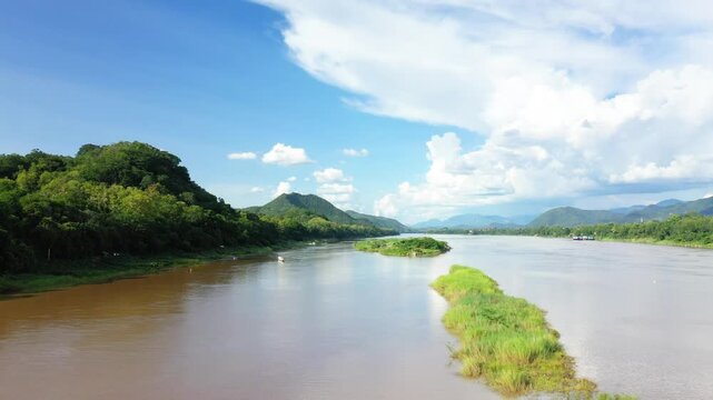 Wide tranquil river flowing through lush green hills under a bright blue sky in Luang Prabang, Laos. Peaceful natural scenery with distant mountains and vibrant clouds.