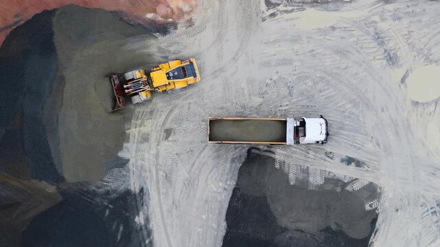 Aerial view of a yellow loader filling a white truck with material on a construction site, contrasting textures and tones, Plouasne, Brittany, France.