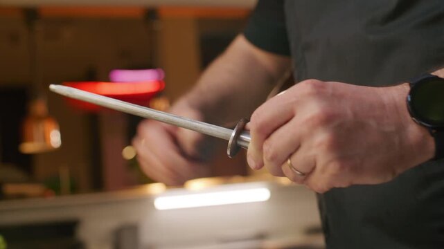 Close up view of chef hands sharpening kitchen knife with honing steel while preparing tools for cooking food in professional kitchen