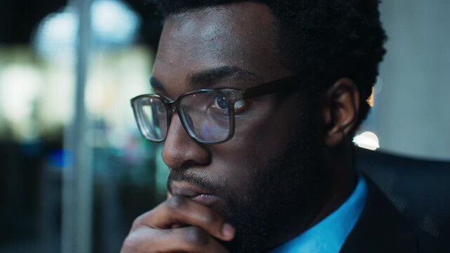 Close portrait of African American businessman thinking at office desk. Adult male wearing glasses resting chin on hand. Watching market screen indoors at night. Making trading decisions after hours.