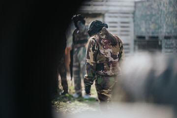 Fototapeta premium A person in camo gear stands with their back to the camera in a paintball arena. Outdoors, players prepare behind barriers in a tactical sport scene.