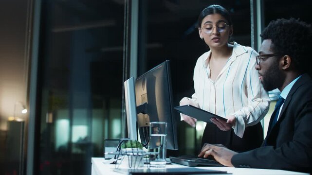 Wide office scene of businesswoman standing by computer desk at night. Adult woman explaining data on tablet. Male colleague listening at monitor. Discussing stock charts indoors after work hours.