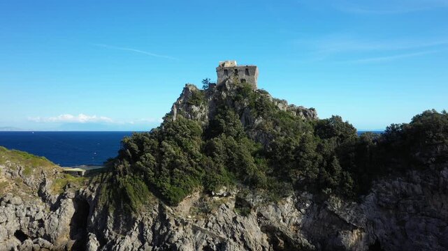 Historic stone watchtower perched on a rocky cliff above the deep blue sea at Capo di Conca, Amalfi Coast, Italy. Bright clear sky and Mediterranean landscape create a dramatic coastal scene.