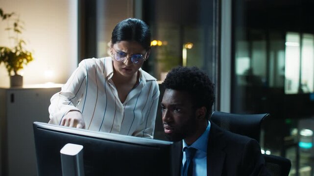 Night office scene of businesswoman pointing at computer screen. Adult female identifying mistake on monitor. African American businessman watching data. Reviewing analytics indoors after hours.