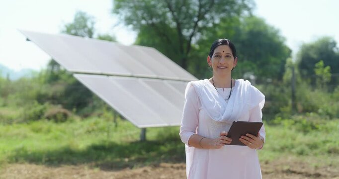 Indian adult elder older woman wear white dress hold tab visit village field work look camera outdoor rural area. Happy old aged female lady staff staring cam do casual pose enjoy day at ground place