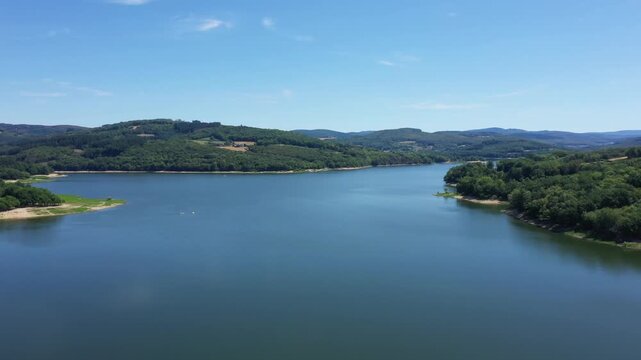 Wide aerial view of the tranquil Panneciere Chaumard reservoir surrounded by lush green hills and forest under a clear blue sky in Burgundy, France.