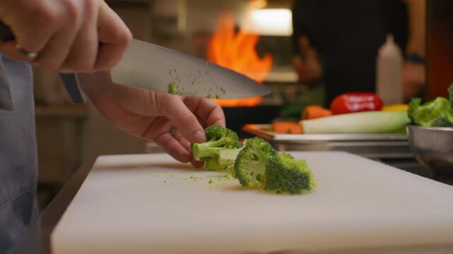 Close up view of chef hands slicing fresh broccoli with knife on cutting board in restaurant kitchen while his colleague cooking flambe with flame in background