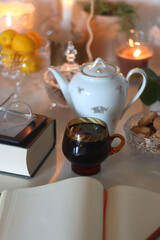 Obraz premium Cup of tea or coffee, teapot, book, notebook, e-reader, glasses, cookies, nuts, lemons, house plants and lit candles on the table. Hygge at home. Selective focus.
