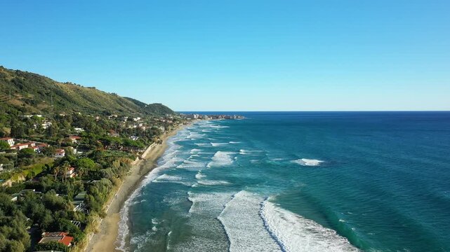 Scenic aerial view of the coastline and turquoise sea in Acciaroli, Italy, with waves gently breaking along the shore and lush green hills in the background.
