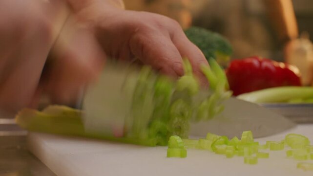 Hands of unrecognizable cook slicing fresh celery into thin pieces with sharp knife on cutting board while preparing vegetable ingredients in restaurant kitchen. Close-up view