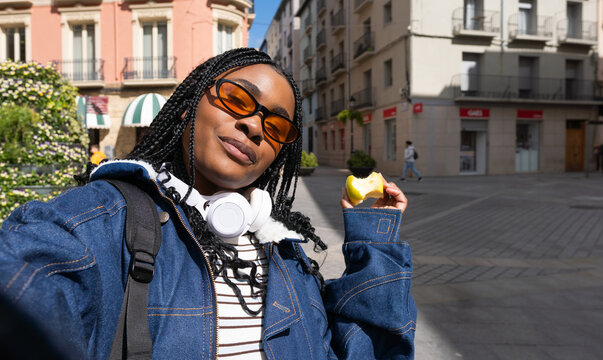 Young black woman eating apple and taking selfie in city center