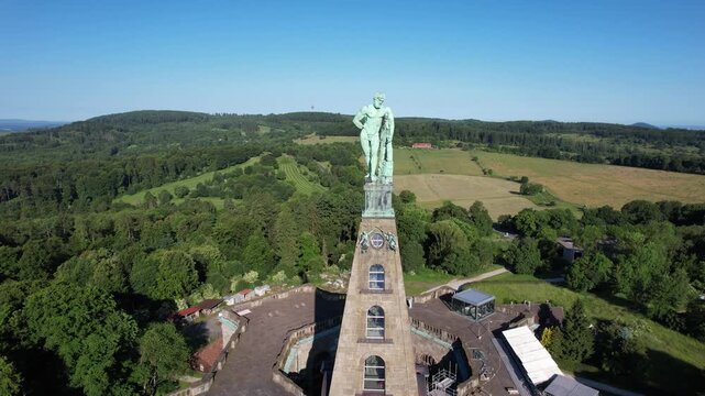 Aerial view of the Hercules Monument standing tall amidst lush greenery, the statue gleaming against the bright blue sky, Kassel, Hessen, Germany.