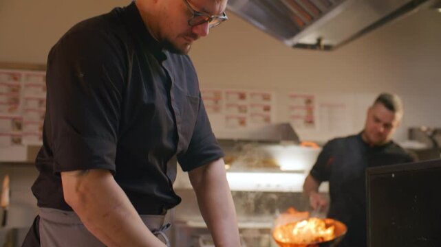 Chef slicing carrot on cutting board with knife while working in professional restaurant kitchen, his colleague performing flambe in background. Tilt-up shot
