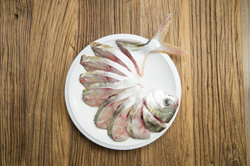 On a round white plate, a fish is expertly sliced and fanned while keeping the head and tail attached. The silver skin and pale flesh show clean knife work on a rustic wooden surface, highlighting pre