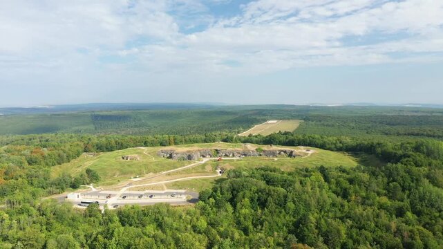 Wide aerial shot of Fort de Douaumont surrounded by lush green forest under a partly cloudy sky in France. The historic fort is set amidst rolling hills and expansive woodland.