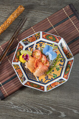 Salmon sashimi curls with cherry tomato, lime, and edible blossoms rest in an ornate patterned plate. Bamboo mat and chopsticks frame the fresh, colorful presentation from a top view.