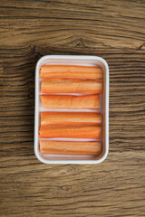A compact white tray displays rows of orange surimi sticks. The simple top view and rustic wood background emphasize neat preparation of a convenient seafood ingredient.