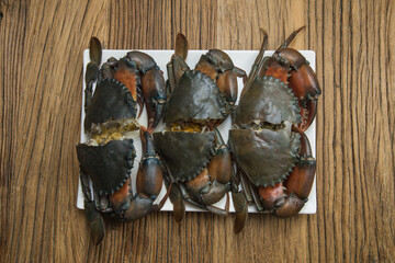 A white tray holds several halved raw mud crabs with shells and claws intact. Set on rustic wood, the arrangement highlights natural color, roe, and ocean freshness ready for seafood cooking.