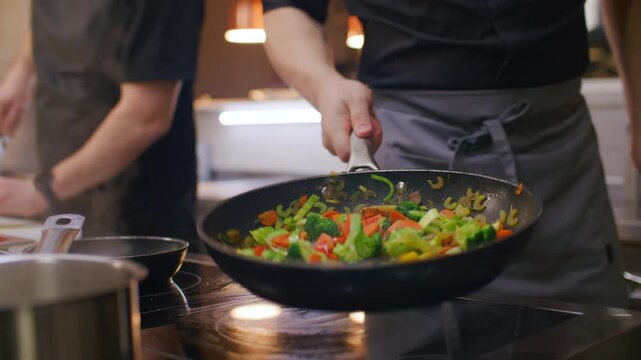 Restaurant chef in apron adding salt to mixed vegetables in frying pan, then tossing ingredients during stir fry cooking on hot stove in kitchen. Close-up view