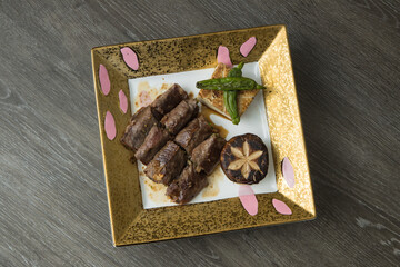 On a decorative square plate, seared beef slices are arranged beside a charred green pepper, a grilled shiitake and a crisp tofu wedge. The clean overhead styling suggests modern fusion dining.