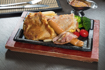 A hot plate set features a crispy pork chop, sausage and bacon with fries, broccoli and cherry tomato. Served on cast iron over a wooden base with a small gravy boat on the side.
