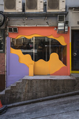 A colorful storefront with curved window graphics and the text "HEI RICE" stands on a sloped street. Air conditioner units and concrete steps frame the small shop entrance in natural daylight.
