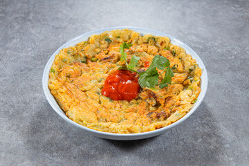 A flat oyster omelette fills a shallow bowl, topped with a spoon of ketchup and fresh cilantro. The egg is speckled with greens and oysters for a homely comfort dish.