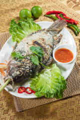 A whole fish with a crust of salt lies on a white platter with lettuce, cilantro and sliced chili. A lemongrass stalk scents the fish, served with a small bowl of bright dipping sauce.